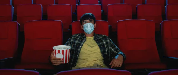 Young guy in casual clothes wearing safety mask, holding popcorn basket while watching movie, visiting cinema alone during coronavirus pandemic — Stock Photo, Image