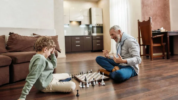 賢いゲームだ。家のリビングルームの床に一緒に座っている間におじいちゃんとチェスをしている小さな男の子 — ストック写真