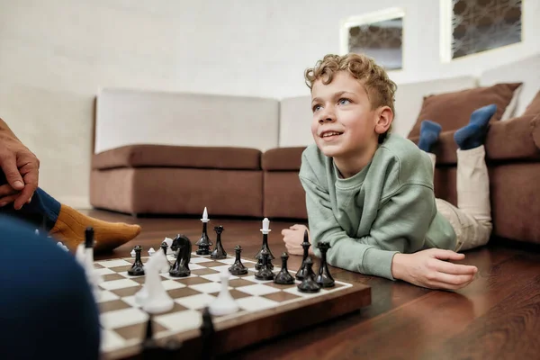 リビングルームで床に横たわっている間に祖父とチェスをしてかわいい男の子,家で一緒に時間を過ごす — ストック写真