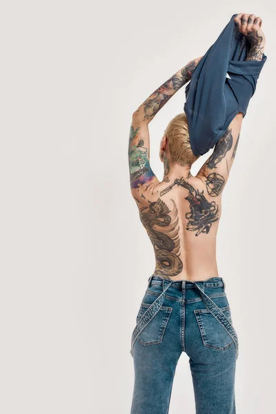 Tattoo and piercing. A pierced and tattooed white girl wearing denim overall standing with her back to a camera finishing taking off a blue t-shirt