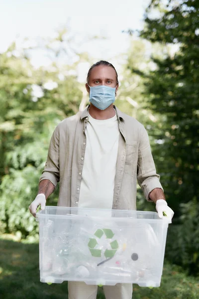 Reciclaje y COVID19. Joven eco activista masculino con mascarilla protectora con papelera de reciclaje mirando a la cámara mientras recoge residuos plásticos en el bosque durante la pandemia de coronavirus — Foto de Stock