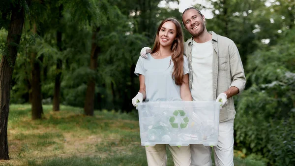 Öko-Familie. Junges fröhliches Paar hält Papierkorb in der Hand und lächelt in die Kamera, während es gemeinsam grünen Wald von Plastikmüll befreit — Stockfoto