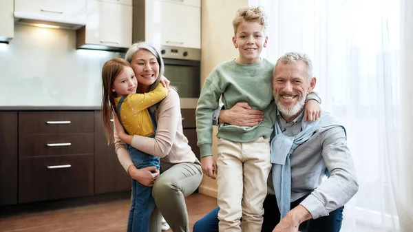 Vrolijke familie. Grootouder met kleinkinderen die plezier hebben en elkaar knuffelen in de eetkamer of keuken thuis, poseren voor foto en glimlachen naar de camera — Stockfoto
