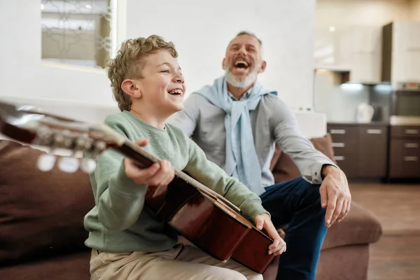 Genç ve yaşlı nesiller arasındaki ilişkiler. Mutlu büyükbaba ve heyecanlı küçük torunu kanepede oturup gülüyor ve gitar çalmaktan zevk alıyorlar. — Stok fotoğraf