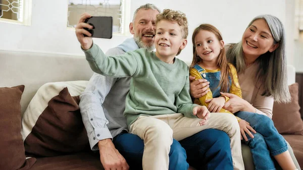 Prachtige momenten. Gelukkig grootouder met twee schattige kinderen kleinkinderen maken van een selfie op de smartphone, ontspannen samen op de bank in de woonkamer — Stockfoto