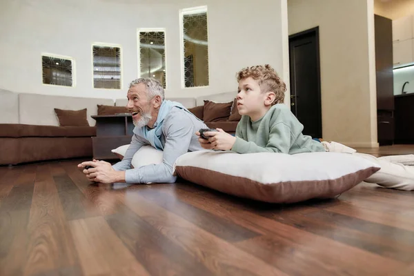 Online competitie. Zijaanzicht van een grootvader met kleinzoon liggend op kussens op de vloer en videospelletjes spelend, tijd samen thuis doorbrengend — Stockfoto