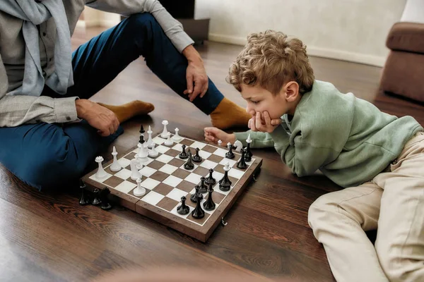 リビングルームの床に一緒に座っている間、かわいい男の子は祖父とチェスをして、家で時間を過ごす。子供のための知的ゲームと活動 — ストック写真