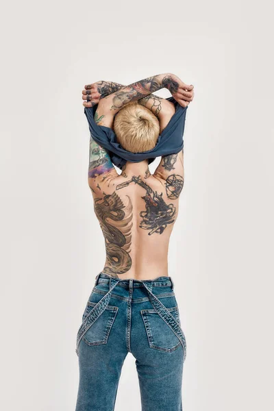 纹身和穿孔。一个全身穿着斜纹斜纹棉布的被纹身穿孔的白人女孩，背对着相机，正在脱下一件蓝色T恤 — 图库照片