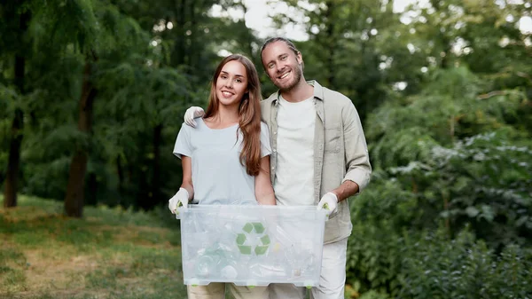 Save Earth from plastic pollution. Young cheerful couple holding recycle bin and smiling at camera while cleaning together green forest from plastic waste — Stock Photo, Image