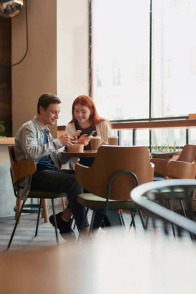 Ένα ζευγάρι εφήβων που διασκεδάζουν βλέποντας κάτι, χρησιμοποιούν τα τηλέφωνά τους, κάθονται σε ένα καφέ μαζί την ημέρα — Φωτογραφία Αρχείου