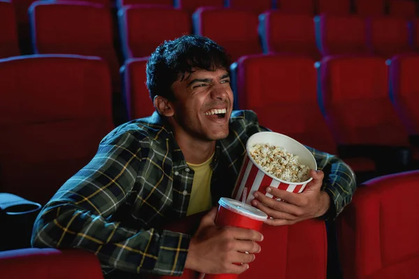 Portrét veselého mladíka, jak se směje, drží v ruce koš na pití a popcorn, zatímco se sám dívá na film v prázdném hledišti divadla — Stock fotografie