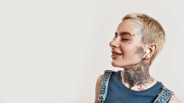 Escuchando música. Un primer plano de una chica blanca tatuada con piercing vistiendo denim en general de pie y escuchando música con sus auriculares inalámbricos ojos cerrados y sonriendo — Foto de Stock