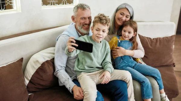 Glückliche Großeltern mit zwei Enkelkindern machen ein Selfie auf dem Smartphone, während sie es sich gemeinsam auf dem Sofa im Wohnzimmer gemütlich machen, sich umarmen und Spaß haben — Stockfoto