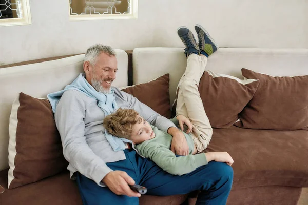 Amoroso avô. Retrato de um avô feliz e neto animado abraçando, se divertindo e brincando enquanto relaxa em um sofá em casa — Fotografia de Stock