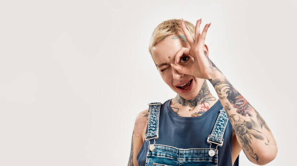 Emotions and postures. A white pierced and tattooed woman showing an OK sign in front of her eye with her mouth open smiling and looking into a camera wearing a denim overall