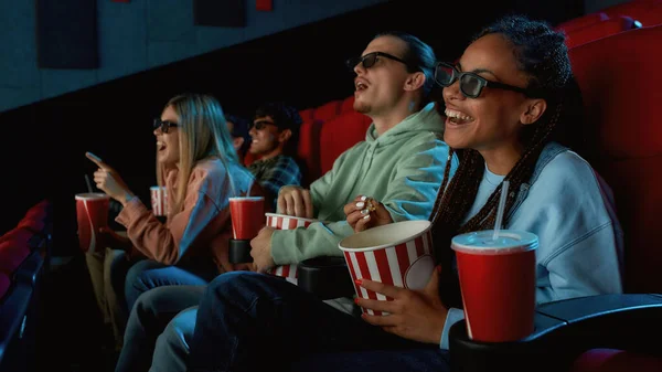 Happy young woman wearing glasses, eating popcorn while watching movie in cinema auditorium together with her friends