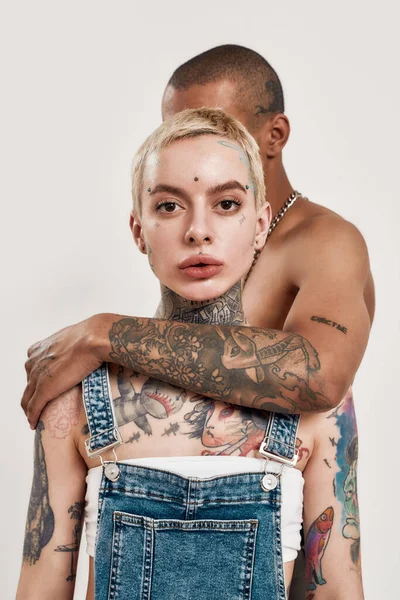 Multikulturellt förhållande. En mörkhyad topless tatuerad kille står bakom en vit piercad tatuerad vuxen flicka kramar henne från en rygg med handen medan hon tittar in i en kamera — Stockfoto