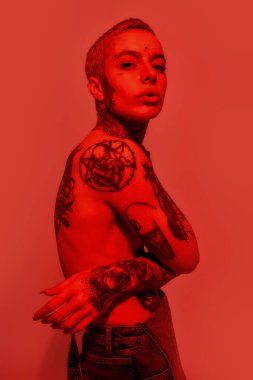 Dövme ve piercing. Dövmeleri ve piercingleri olan beyaz bir kadın elleri göbeğinde, göğüslerini kapatan bir kameraya bakıyor.