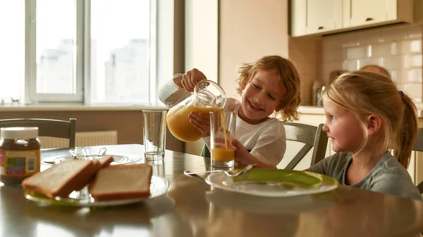 Küçük kız ve erkek kardeş sabahleyin evde kendi başlarına kahvaltı yapıyorlar. Neşeli çocuk gülümserken bardaklara portakal suyu dökerken — Stok fotoğraf