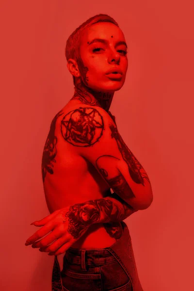 Tatuagem e piercing. Uma mulher branca com tatuagens e piercing em pé topless lateralmente olhando para uma câmera com as mãos cruzadas em seus seios barriga cobertura — Fotografia de Stock