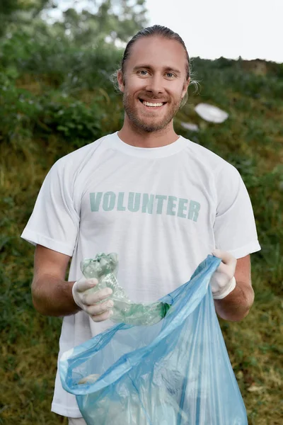 リサイクルの概念。笑顔の青年、制服を着た男性ボランティア、森や公園でプラスチックごみを集めるゴム手袋の垂直ショット — ストック写真