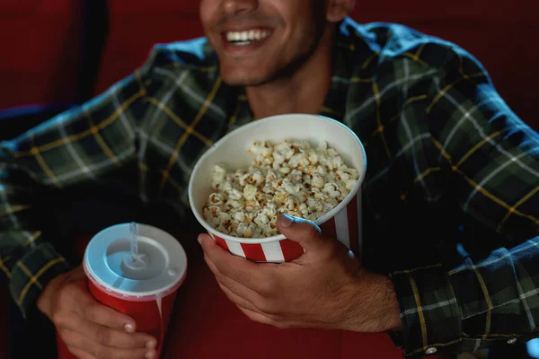 Gesneden portret van vrolijke jonge man glimlachend, met een drankje en popcorn mand, terwijl het kijken naar de film alleen in lege theater auditorium — Stockfoto