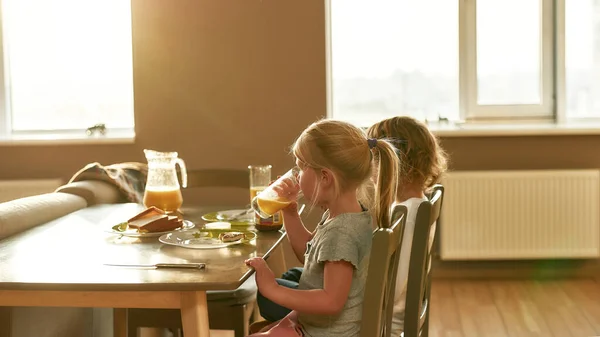 Lillebror och syster dricker apelsinjuice, äter frukost eller lunch medan de sitter vid bordet i köket — Stockfoto