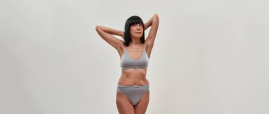 Olgun model. Çekici yarı çıplak, beyaz, son sınıf öğrencisi, kollarını kaldırmış, başını çevirmiş, gri arka planda iç çamaşırlarıyla poz veriyor.