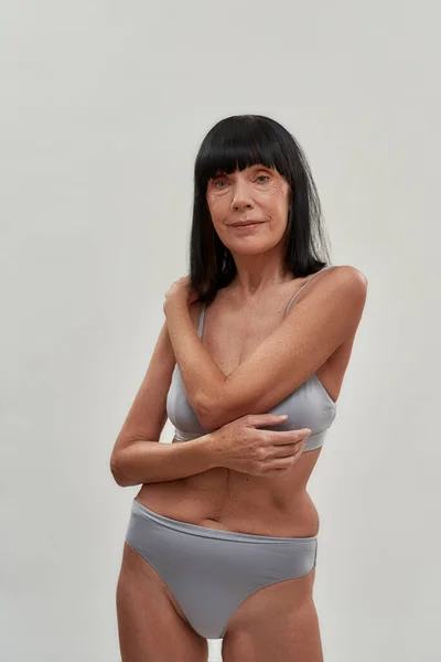 Human age. Portrait of a beautiful half naked caucasian senior woman in lingerie posing against grey background