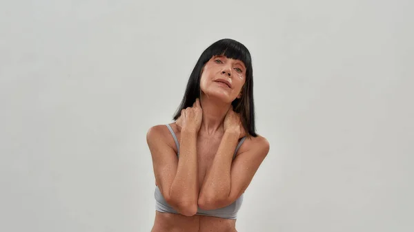 Belle moitié nue femme caucasienne d'âge moyen touchant son cou, regardant la caméra tout en posant en lingerie en studio, debout sur fond gris — Photo