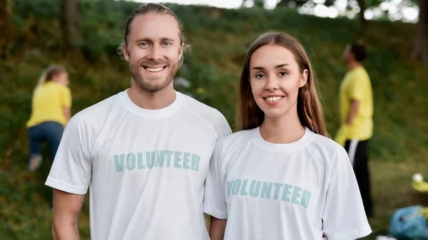 Charity work. Portrait of two happy eco activists, man and woman wearing uniform smiling at camera while cleaning forest or park with group of volunteers
