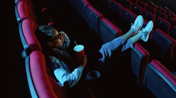 Stijlvolle jonge vrouw in 3D bril zet haar voeten op een stoel, het vasthouden van een drankje tijdens het kijken naar de film alleen in lege theater auditorium — Stockfoto