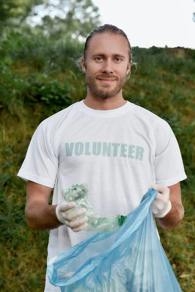 片付けなさい。笑顔の青年、制服を着た男性ボランティア、森や公園でプラスチックごみを集めるゴム手袋の垂直ショット — ストック写真