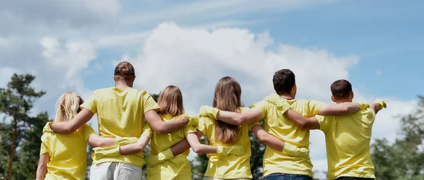 Любите свою натуру. Группа молодых добровольцев в форме и резиновых перчатках обнимаются и смотрят на зеленый лес перед ними, вид сзади — стоковое фото