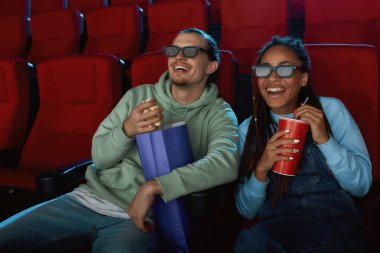 3D gözlük takan neşeli genç çift sinemada film izlerken gülümsüyor, patlamış mısır yiyor ve gazoz içiyor.