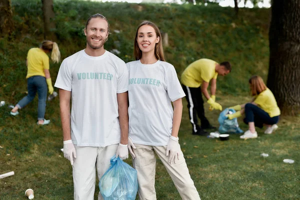 Заботиться о природе. Два счастливых эко-активиста в униформе и резиновых перчатках улыбаются в камеру, очищая лес от пластиковых отходов, мусор с группой добровольцев — стоковое фото