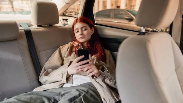 Nastolatka w słuchawkach za pomocą smartfona podczas siedzenia na tylnym siedzeniu w samochodzie — Zdjęcie stockowe