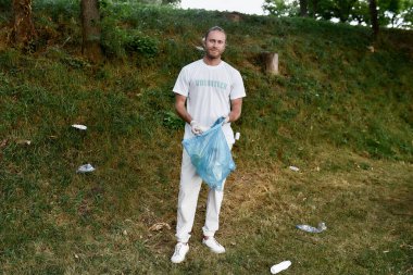 Dünyayı kurtar. Üniformalı ve plastik eldivenli genç bir çevreci orman veya park temizlerken çöp ve çöp torbası tutuyor.
