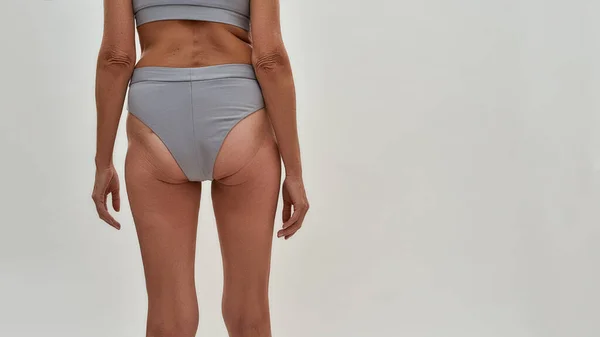 Visão traseira de uma mulher sênior magro seminua em lingerie em pé contra fundo cinza no estúdio — Fotografia de Stock