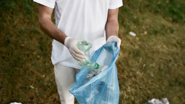 Young male volunteer wearing uniform and rubber gloves collecting plastic waste in the forest or park, cropped shot — Stock Photo, Image