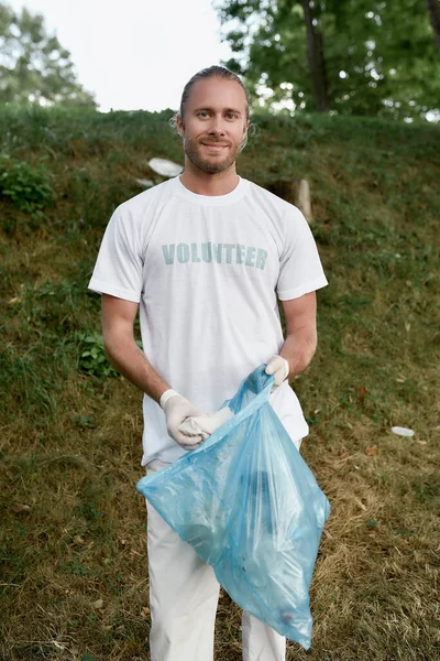 Eco activista. Joven hombre sonriente con uniforme y guantes de goma sosteniendo bolsa de plástico con basura mientras limpia el bosque o el parque, mirando a la cámara — Foto de Stock