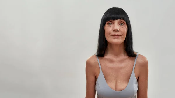 Retrato de una hermosa mujer mayor mirando a la cámara mientras está de pie sobre un fondo gris, posando medio desnuda en el estudio —  Fotos de Stock