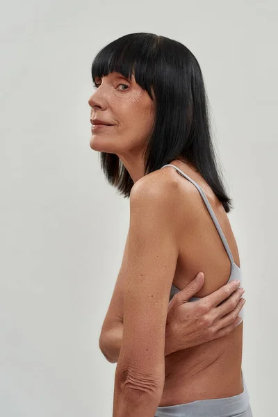 Beleza madura natural. Vertical tiro de uma mulher caucasiana sênior em lingerie abraçando-se, olhando para a câmera enquanto posando isolado sobre fundo cinza — Fotografia de Stock