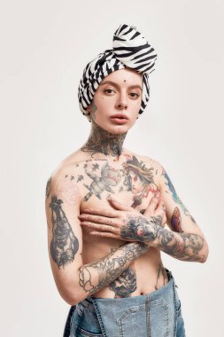 Dövme ve piercing. Beyaz piercingli ve dövmeli bir kadın göğüslerini saran bir türbanla duruyor göğsünde bir kot pantolon var ve kameraya bakıyor.