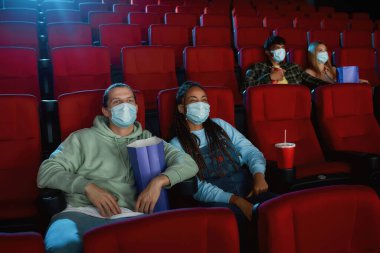 Sinema salonunda film izlerken virüs hastalığından korunmak için maske takan neşeli genç bir çift.