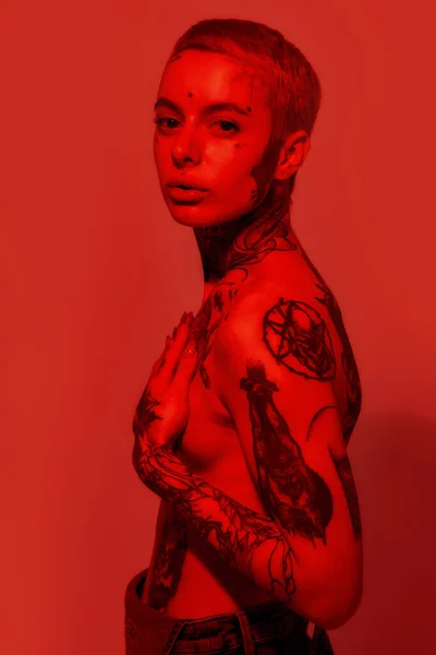 Tatuagem e piercing. Uma mulher branca com tatuagens e piercing em pé topless lateralmente olhando para uma câmera com as mãos cobrindo seios — Fotografia de Stock