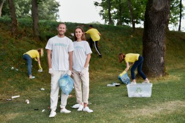Üniformalı ve lastik eldivenli iki mutlu arkadaş plastik atıktan ormanı temizlerken kameraya gülümseyen eko aktivist, bir grup gönüllüyle çöpler.