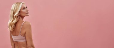 Panoramik pankart. Güzel, yarı çıplak, orta yaşlı sarışın kadın pembe arka planda poz verirken başka tarafa bakıyor, mesaj için boşluk kopyala