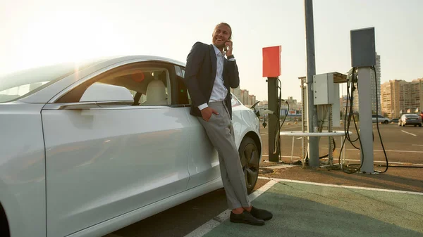 Happy man praten over de telefoon terwijl elektrische auto opladen — Stockfoto