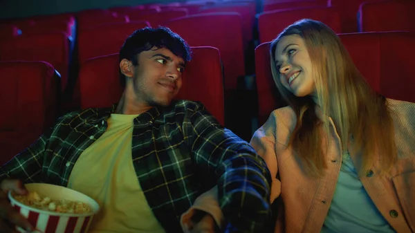 Привлекательная пара, молодой человек и женщина, улыбающиеся друг другу, держащиеся за руки во время просмотра фильма, сидящие в кинотеатре, имеющие романтическое свидание в кино — стоковое фото